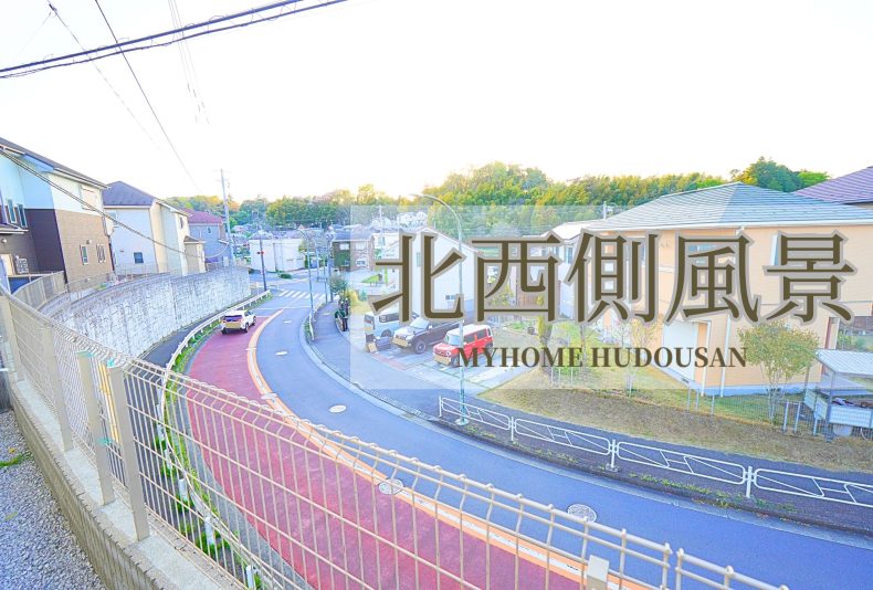 実際の物件敷地内から北西側の風景を写した写真。綺麗な歩道が付いた幅広の道路が眼下に流れる開放的な景色。周囲に緑が多く、明るい住宅街の風景の中に建つ物件である事がわかる画像。

